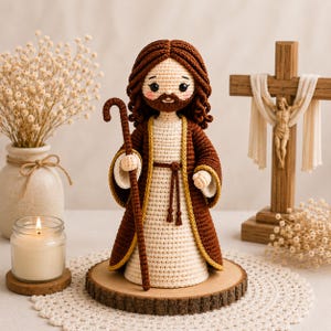 May include: A crocheted doll depicting a figure with brown hair and a beard, wearing a cream-colored robe with a brown outer garment and a tied belt. The figure holds a brown shepherd's crook and stands on a circular wooden base.