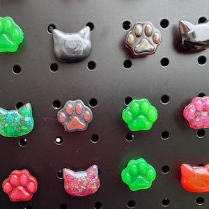 May include: A collection of twelve small resin charms shaped like cat heads and paw prints in various colors including green, black, red, and pink. Some charms feature glitter or metallic finishes, all arranged on a black pegboard display.