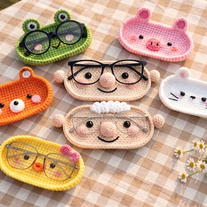 Puede incluir: Una colección de siete bandejas ovaladas de ganchillo diseñadas para guardar gafas. Las bandejas presentan varios rostros de animales y personajes, incluyendo una rana verde, un cerdo rosa, un oso naranja, un gato blanco y dos figuras humanas.