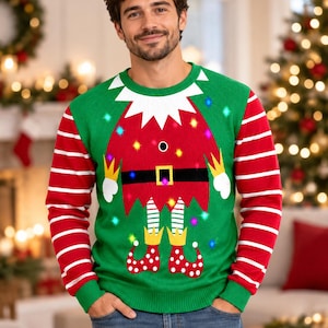 Peut inclure: Un pull vert à manches longues orné d'un motif de tunique d'elfe rouge avec une ceinture noire, un col blanc et des manches rayées. Le design intègre des lumières LED colorées dans le tricot, accompagnées de chaussures d'elfe à pois rouges et blancs.