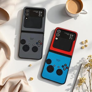 May include: Two foldable smartphones featuring custom skins designed to resemble retro handheld game controllers. One device has a grey base with black buttons, while the other has a blue base with black buttons and a red top section around the camera.