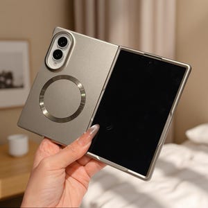 May include: A metallic champagne-colored foldable smartphone case featuring a dual-camera cutout at the top left and a circular magnetic charging ring centered on the back panel. The case is shown in an open position with a black screen visible.