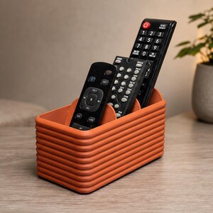 May include: A rectangular, terracotta-colored storage organizer with a ribbed, horizontal texture. The container features three internal compartments, currently holding three black remote controls of varying sizes and button configurations.