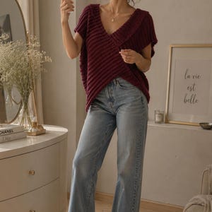 Könnte beinhalten: Ein dunkelrotes, kurzärmeliges Strickoberteil mit V-Ausschnitt und gerippter Textur, kombiniert mit einer hellblauen Jeans. Das Oberteil hat ein Wickeldesign auf der Vorderseite mit kurzen Ärmeln und einer lockeren Passform, getragen mit hellen Sandalen.