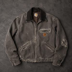 May include: A distressed charcoal grey canvas jacket featuring a dark brown corduroy collar, a full-length front zip, and a plaid interior lining. The garment includes a zipped chest pocket with a Carhartt logo patch and two lower pockets.