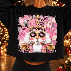 Puede incluir: Una sudadera negra de manga larga con el gráfico de un cascanueces con estampado de leopardo rosa, charreteras doradas y gafas de sol en forma de corazón, sobre un fondo de manchas de acuarela rosa, lazos y adornos.
