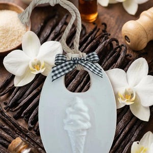 May include: A white oval ceramic hanging ornament featuring a raised ice cream cone relief in the center. The ornament includes a beige rope loop for hanging and is decorated with a small black and white gingham ribbon bow at the top.