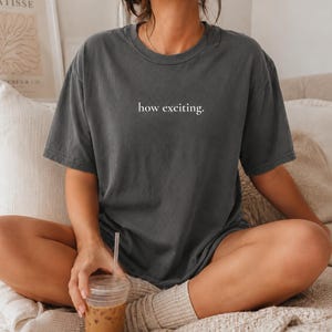 Peut inclure: Un t-shirt à manches courtes gris anthracite avec le texte "how exciting." imprimé en police serif blanche au centre de la poitrine. Le vêtement présente une encolure ronde et une coupe décontractée avec un aspect légèrement délavé.