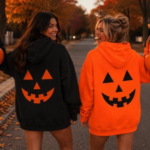 Puede incluir: Una sudadera con capucha negra con un estampado de cara de calabaza naranja en la espalda y una sudadera con capucha naranja brillante con un estampado de cara de calabaza negra en la espalda, cada una con un cubo de plástico naranja de calabaza.