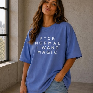 May include: A periwinkle blue short-sleeve t-shirt featuring the white printed text F*CK NORMAL I WANT MAGIC on the chest. The garment has a crew neckline and a loose, relaxed fit, paired with light blue denim shorts with a frayed hemline.
