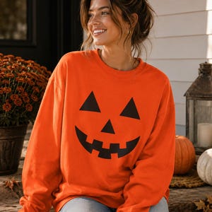 May include: A bright orange long-sleeve sweatshirt featuring a large black graphic of a jack-o'-lantern face with two triangular eyes, a triangular nose, and a wide, jagged, toothy grin printed on the center of the chest area.