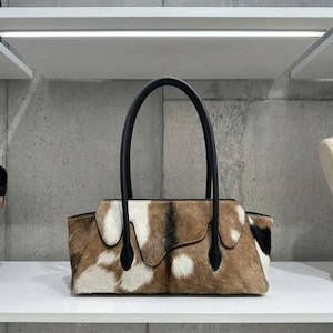 May include: A rectangular handbag featuring a textured cowhide exterior with a mottled pattern of brown, white, and black patches. The bag includes two tall, smooth black leather handles attached to the top, with a curved flap overlay design.