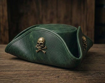 Op de afbeelding: Een donkergroene driekante hoed met een verweerde textuur en twee metalen, messingkleurige schedel- en gekruiste beenderen-emblemen bevestigd aan de omhoogstaande zijkanten. De hoed heeft een gestructureerde kroon en brede, omgevouwen randen.