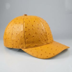 Puede incluir: Una gorra de béisbol color amarillo mostaza con un patrón texturizado de piel de avestruz. La gorra está construida con seis paneles, un botón a juego en la parte superior central y una visera curva, con una textura punteada uniforme.