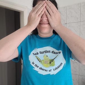 Peut inclure: Un t-shirt bleu turquoise à manches courtes avec un motif central représentant une tortue de mer verte. Le design comprend le texte "Sea turtles dance in the shores of Lebanon" imprimé en lettres noires autour de l'illustration.