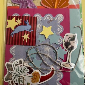 May include: A collection of assorted paper craft ephemera including a purple glitter starfish, an orange seashell, a red square with a blue rabbit, yellow and blue stars, a cocktail glass cutout, and a floral sprig, all secured with a silver paperclip.