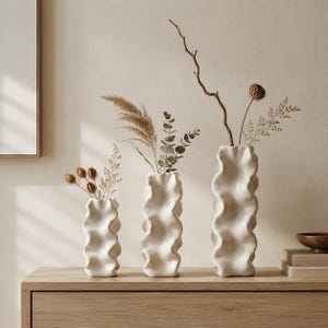 May include: Three white ceramic vases of varying heights, each featuring a distinct wavy, rippled texture. The vases are filled with dried botanical stems, including pampas grass, eucalyptus leaves, and thin, bare branches with small seed pods.