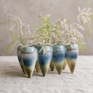 May include: A multi-chambered ceramic vase with a gradient glaze transitioning from a pale yellow base to a deep blue midsection and a turquoise rim, containing several sprigs of delicate white feathery flowers with thin green stems.