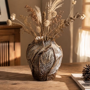 May include: A dark brown ceramic vase with a textured, leaf-like relief pattern and a distressed white finish, filled with an arrangement of dried beige grasses, fluffy seed heads, and eucalyptus stems, creating a natural and earthy display.