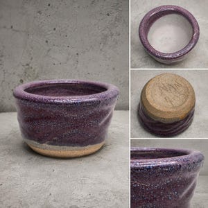 Può includere: Una ciotola in ceramica rotonda con uno smalto viola maculato all'esterno e un interno bianco liscio. La base non è smaltata, mostrando una consistenza di argilla beige con l'iscrizione incisa a mano NAW 903 visibile sul fondo.