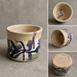 Puede incluir: Una maceta cilíndrica de cerámica con una base de color crema y pinceladas abstractas pintadas a mano en azul oscuro, morado y verde apagado. El recipiente tiene una abertura ancha, un interior liso y una base plana con marca.
