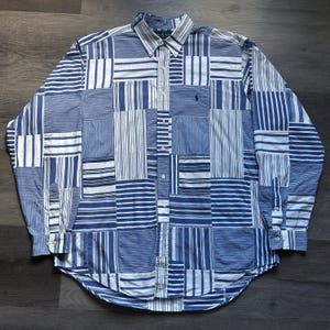 May include: A long-sleeve button-down shirt featuring a patchwork design of various blue and white striped patterns. The shirt has a pointed collar, a front button placket, and a small dark blue embroidered logo on the left chest area.