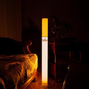 May include: A tall, cylindrical floor lamp designed to resemble a cigarette. The top section is a textured yellow, while the lower portion is white and features the black text Marlboro MEDIUM. The lamp stands on a clear, square base.