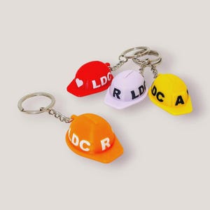Pode incluir: Quatro porta-chaves em miniatura de capacete de segurança nas cores vermelho, roxo claro, amarelo e laranja. Cada capacete de plástico possui um anel e corrente de metal. O capacete vermelho tem um coração branco e o texto LDC, o roxo tem o texto LDC R em preto, o amarelo o texto LDC A em preto e o laranja o texto LDC R em branco.