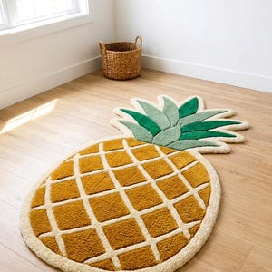 Puede incluir: Una alfombra de mechones con forma de piña con un cuerpo amarillo dorado que presenta un patrón de cuadrícula de color crema. La parte superior tiene hojas verdes puntiagudas en dos tonos y toda la forma está delineada con un borde crema.