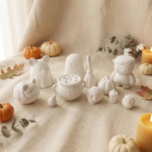 May include: A collection of small white matte ceramic Halloween figurines including a ghost, a mummy, a cauldron, a skull, an owl, a broom, and a Frankenstein-like figure, arranged alongside miniature orange pumpkins and yellow candles.