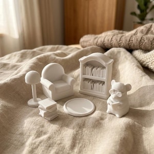 May include: A set of white miniature furniture and a mouse figurine including a rounded armchair, a two-shelf bookcase filled with books, a floor lamp, a stack of four square books, a circular rug, and a mouse wearing glasses holding a book.