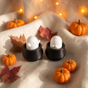Puede incluir: Dos pequeñas figuras de fantasmas blancos, cada una colocada dentro de un caldero negro. Los fantasmas tienen rasgos faciales simples y formas redondeadas. La escena está decorada con varias calabazas naranjas y hojas de otoño.