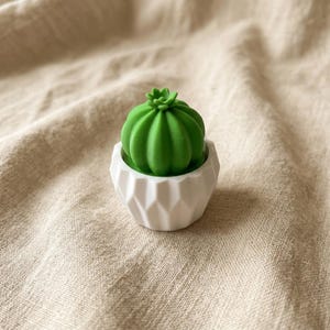 May include: A small, bright green cactus figurine with a rounded, ribbed top and a tiny sprout at the center. The cactus is seated inside a white, geometric, multi-faceted pot that features a series of triangular indentations around its exterior.