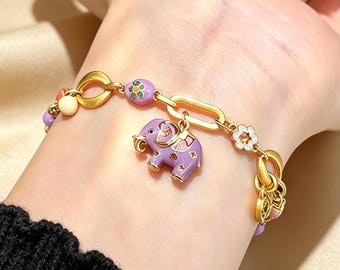 May include: A gold-tone chain bracelet featuring a central purple enamel elephant charm, accented with small purple flower beads, a white flower charm with clear stones, and additional small round beads in various colors along the metal links.