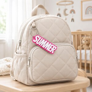 Può includere: Uno zaino trapuntato color beige con manico superiore, due spallacci e una tasca frontale con cerniera. Un portachiavi rettangolare rosa acceso con la scritta bianca "SUMMER" è fissato all'anello metallico sul lato dello zaino.