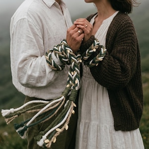 Puede incluir: Un accesorio de cuerda trenzada con cordones entrelazados en blanco, verde oscuro y yute natural, decorado con pequeños dijes metálicos en forma de hoja dorada. La cuerda termina en borlas sueltas y deshilachadas con textura.