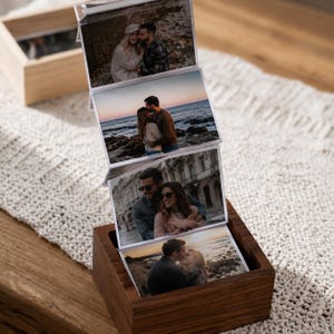 May include: A dark wood square box base holding a vertical accordion-style display of four rectangular photographs. Each photo features a couple in various outdoor and urban settings, including a beach at sunset and a city street scene.