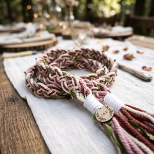 Peut inclure: Une corde tressée composée de fils crème, vieux rose et rouge foncé entrelacés avec de petites feuilles vertes. Les extrémités sont fixées par un fil blanc et portent une étiquette en bois gravée d'une couronne de feuilles et des lettres J&K.