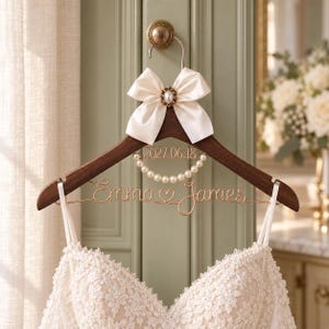 Puede incluir: Una percha de madera oscura con un lazo de satén crema con un detalle de perla, un collar de perlas blancas y letras de alambre en oro rosa que dicen Emma y James con un corazón y la fecha 2027.06.18, sosteniendo un vestido de encaje blanco.