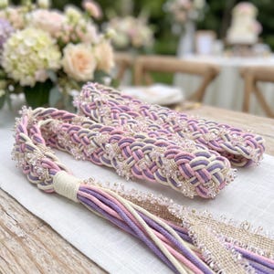 May include: A decorative braided cord featuring interwoven strands of lavender, light pink, and cream-colored fabric. The cord is embellished with clear, faceted beads throughout the weave and terminates in a long, multi-colored fabric tassel.