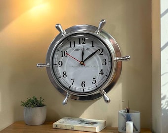 May include: A silver-toned wall clock shaped like a ship's wheel with a white face, black numerals, and black hands. The clock face features the text SHIP'S TIME and HARBOUR NAUTICALS in black lettering, with a red second hand.