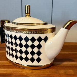 May include: White ceramic teapot with a black and white checkered pattern on the body. The teapot has gold trim around the lid, base, and spout. The lid has a gold handle. A tag is attached to the teapot.