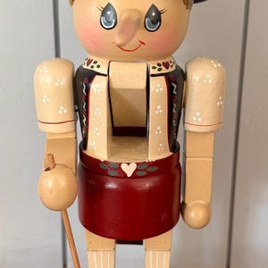 May include: Wooden nutcracker figurine with a painted face, wearing a red hat and skirt, and a black vest with white polka dots. The figurine is holding a wooden stick with a round object.