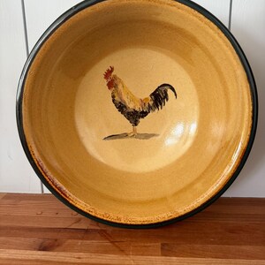 May include: A round, ceramic bowl with a rooster illustration. The bowl has a yellow interior with a black rim. The rooster is depicted in shades of brown, yellow, and black. The bowl is likely used for serving food.