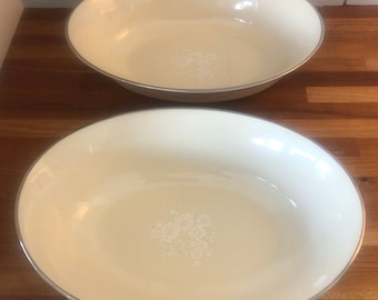 May include: Two white oval ceramic bowls with a silver rim and a floral design. The bowls are on a wooden surface.