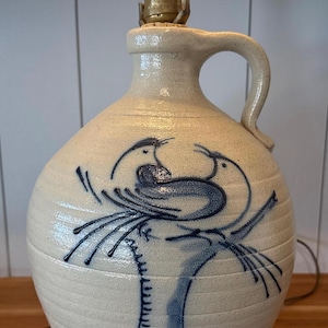 May include: A cream-colored ceramic lamp with a blue bird design. The lamp has a jug shape with a handle and a brass lamp holder. A bright white light bulb is screwed into the lamp.