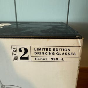 K&ouml;nnte beinhalten: Eine wei&szlig;e Schachtel mit der Aufschrift "SET OF 2 LIMITED EDITION DRINKING GLASSES 399ml". Die Schachtel ist teilweise sichtbar, mit einem Blick auf ein klares Glas.