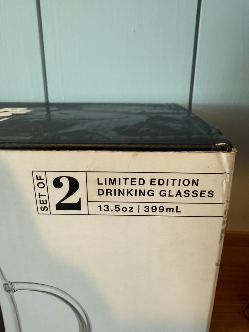 K&ouml;nnte beinhalten: Eine wei&szlig;e Schachtel mit der Aufschrift "SET OF 2 LIMITED EDITION DRINKING GLASSES 399ml". Die Schachtel ist teilweise sichtbar, mit einem Blick auf ein klares Glas.