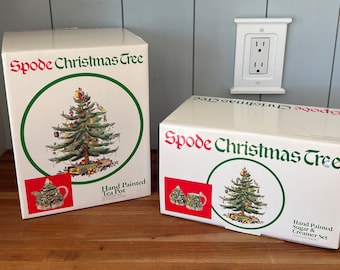 May include: Two boxes of Spode Christmas Tree china. The box on the left is for a hand-painted tea pot. The box on the right is for a hand-painted sugar and creamer set. Both boxes feature a green Christmas tree with red and gold ornaments.