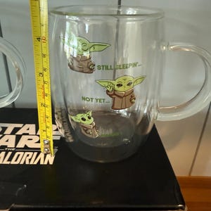 K&ouml;nnte beinhalten: Klarer Glasbecher mit Grogu aus The Mandalorian, mit den Aufschriften "STILL SLEEPIN'" und "NOT YET...". Der Becher ist etwa 10 cm hoch und hat einen Henkel. Der Becher ist Teil eines Sets von zwei Trinkgl&auml;sern in limitierter Auflage.